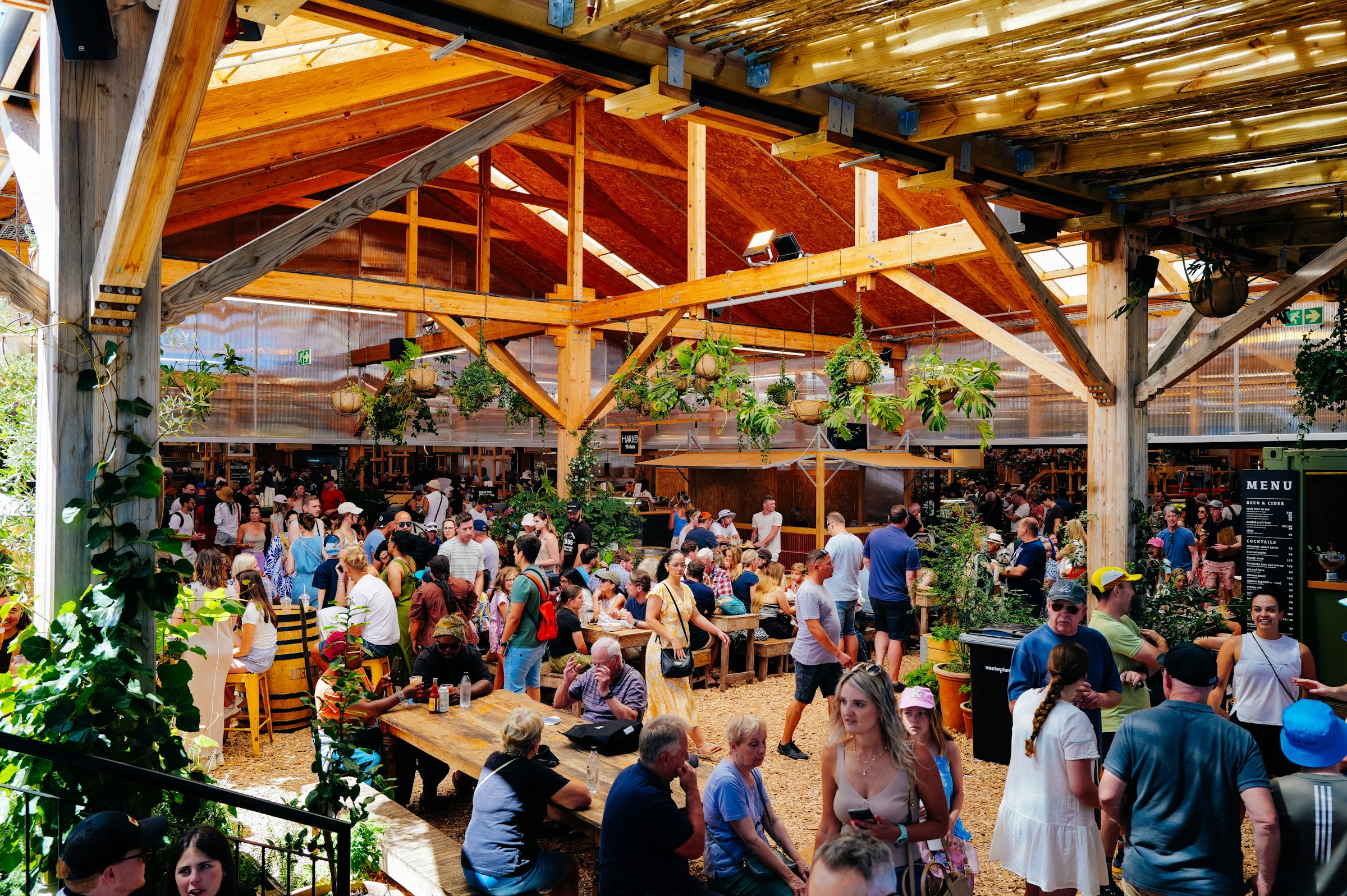 Een volle overdekte biertuin onder houten balken, met mensen aan lange tafels.